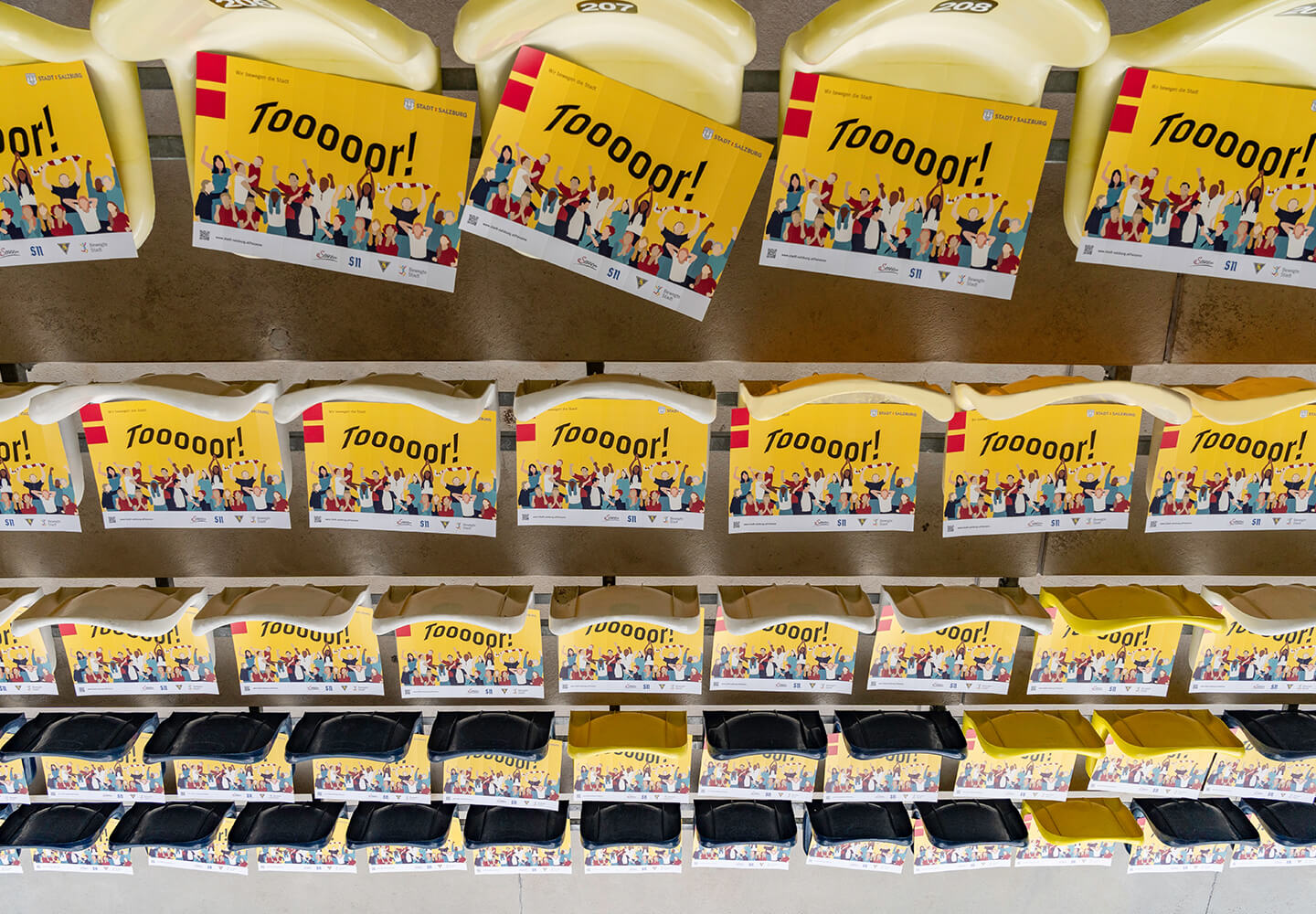 mehrere Faltklatschen mit jubelnden Menschen darauf liegen auf den Sitzen der Tribüne am Fußballplatz